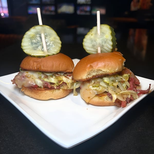 Two sliders with melted cheese, corned beef, and sauerkraut, each topped with a pickle slice.