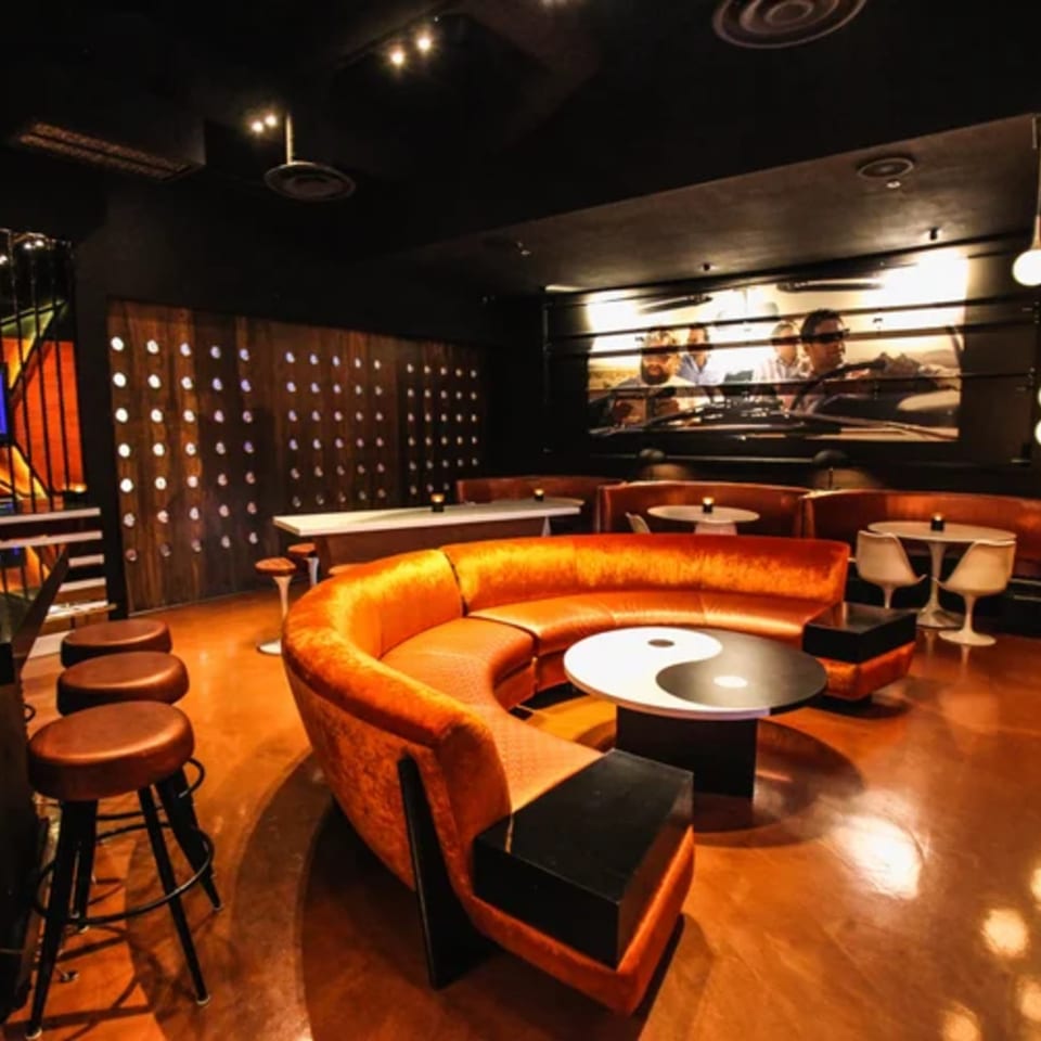 A sports bar interior with an orange curved sofa, yin-yang table, and a large screen.