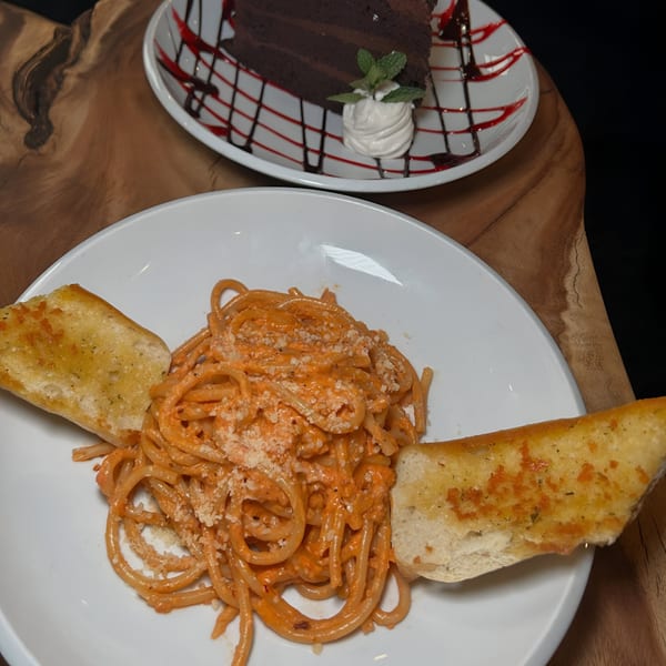 Spaghetti with red sauce and garlic bread, with a slice of chocolate cake in the background.