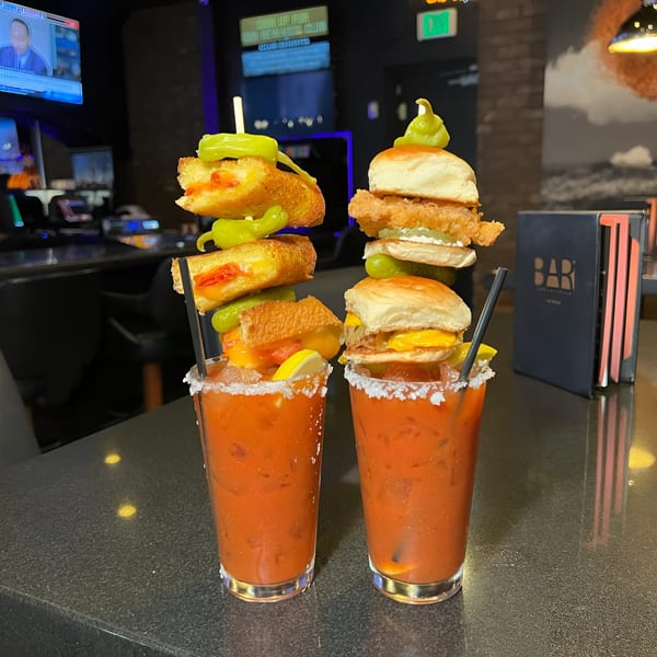 Two Bloody Marys garnished with grilled cheese, sliders, and peppers on a bar top.