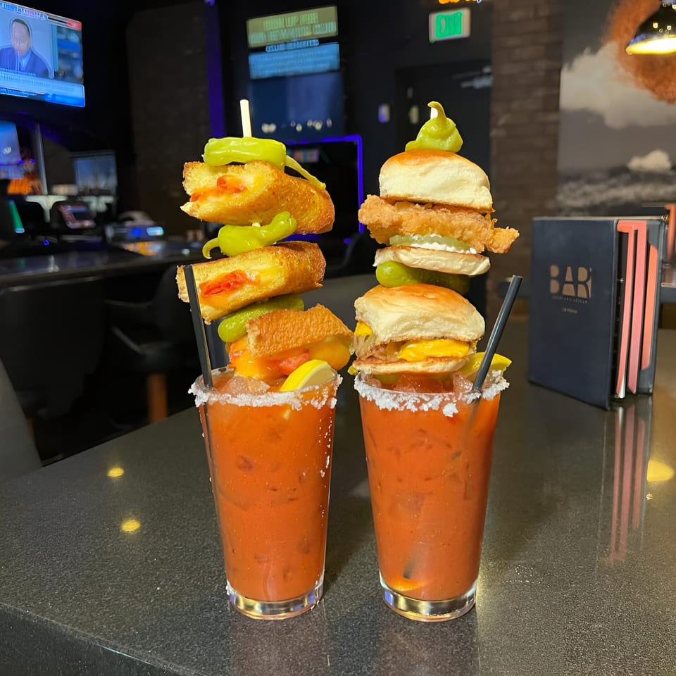 Two Bloody Marys garnished with grilled cheese, sliders, and peppers on a bar top.