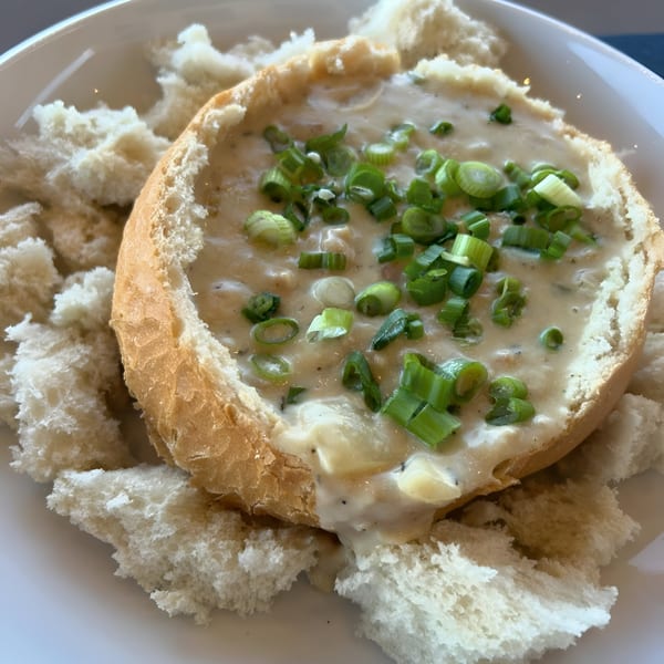 Creamy clam chowder in a bread bowl, garnished with green onions, served with bread pieces.