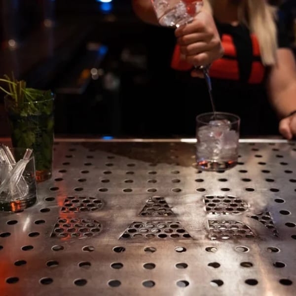 Bartender pouring a drink into a glass with ice on a metal bar top with the Born And Raised logo.