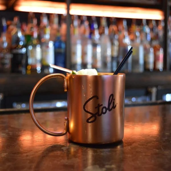 A Stoli-branded copper mug with a drink, lime, and straws on a bar top.